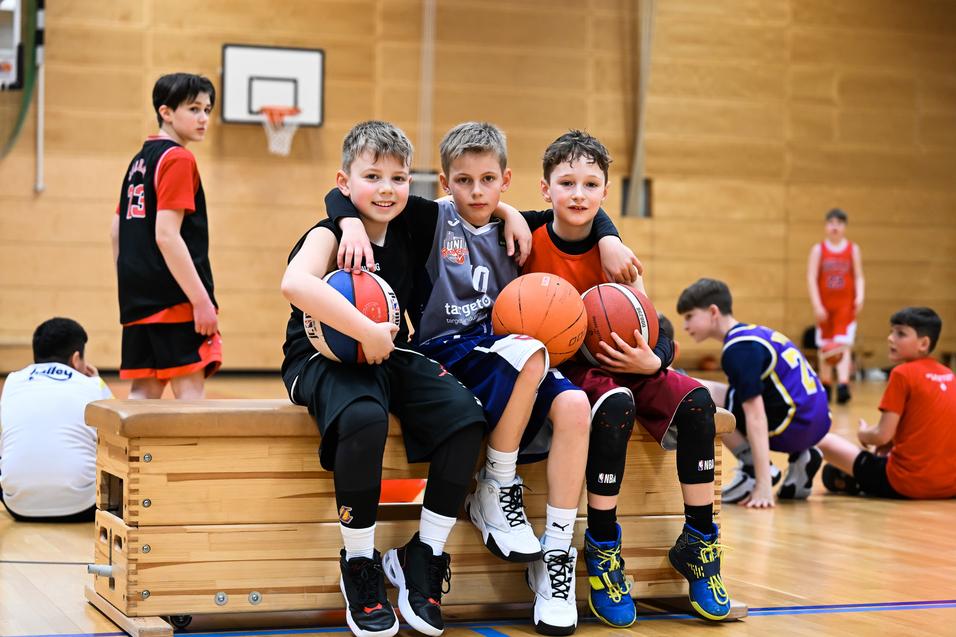 Drei Kinder posieren auf einer Bank in einer Sporthalle, jeder hält einen Basketball. Im Hintergrund weitere Spieler.