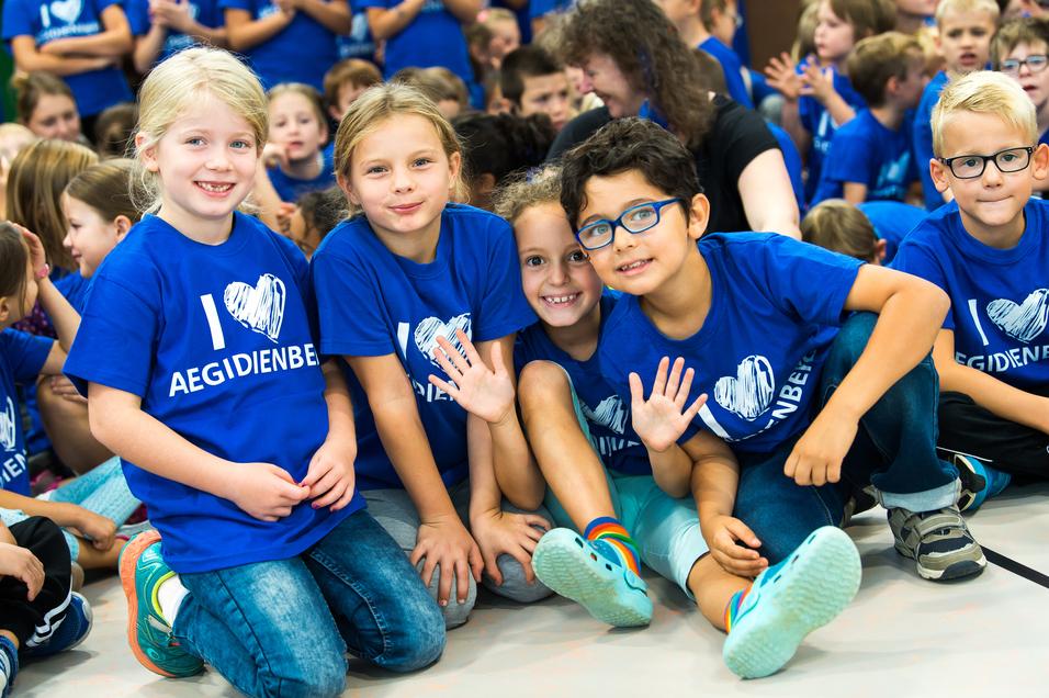 Vier Kinder in blauen T-Shirts mit dem Aufdruck "I ❤️ AEGIDIENBE" posieren lächelnd auf einem Boden.